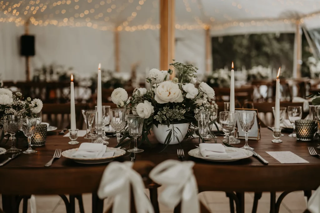 Centre de table floral blanc avec bougies fines allumées et art de la table élégant, sous une tente décorée de guirlandes lumineuses à La Maison Villeneuve. Exemple d’ambiance cohérente illustrant les bonnes pratiques évoquées dans l’article sur les erreurs à éviter en décoration de mariage.