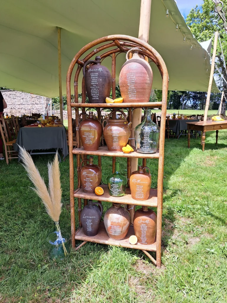 Plan de table réalisé avec des cruches anciennes calligraphiées, présenté sur une étagère en rotin, un exemple original de location déco mariage.