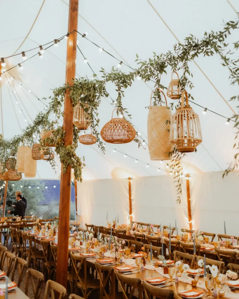 Décoration de dîner de mariage sous tente avec lanternes en rotin, guirlandes lumineuses et tables fleuries pour une ambiance élégante et chaleureuse.