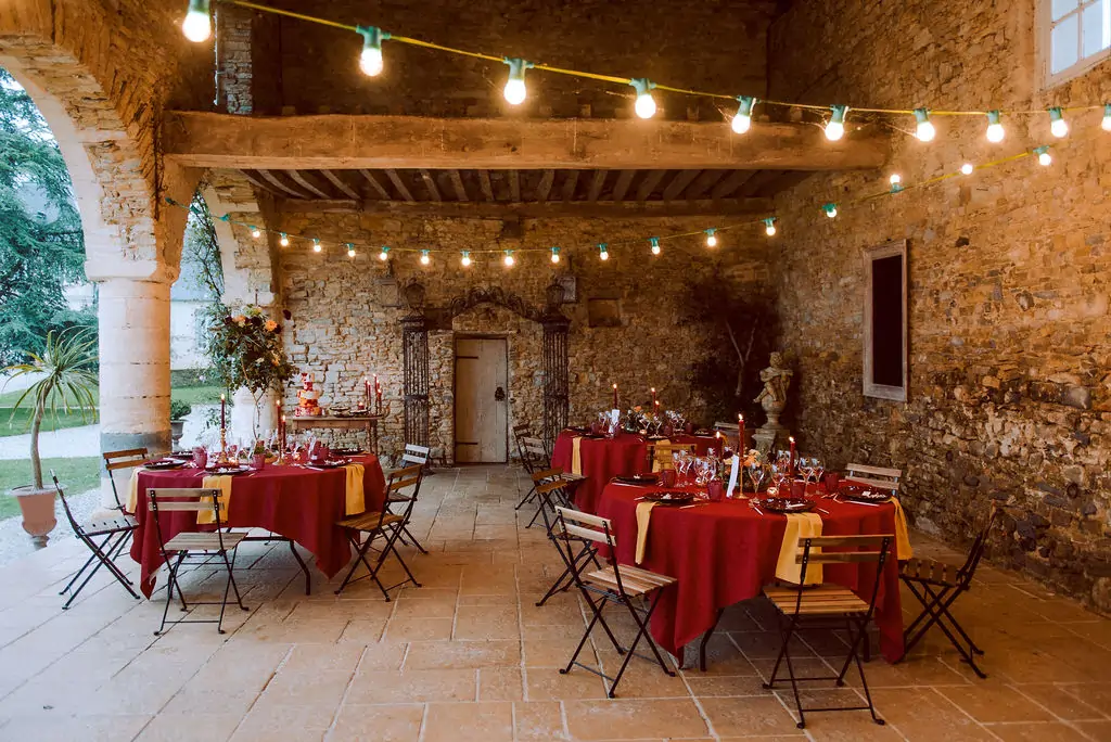 Tables de mariage installées sous une arcade en pierre, éclairées par des guirlandes lumineuses, avec un mobilier pliant naturel et une décoration florale chaleureuse.