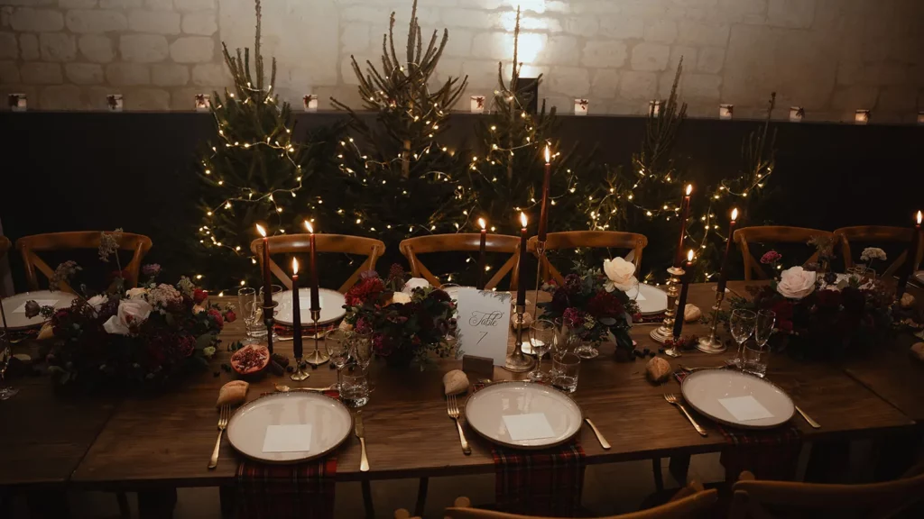 Table de mariage en bois mise en scène avec un mobilier chaleureux, des bougies sombres et des compositions florales d’hiver, devant une rangée de sapins illuminés.
