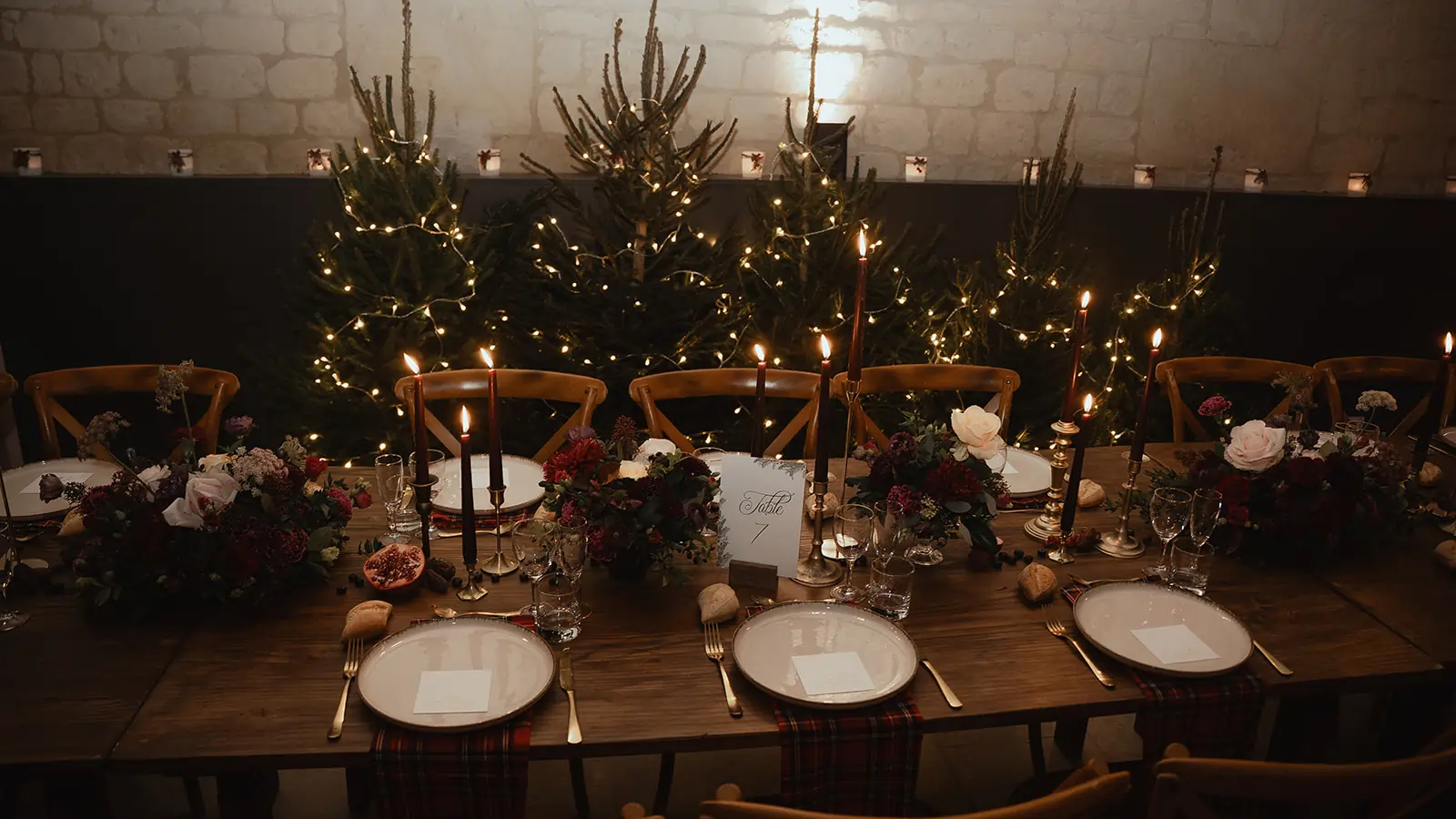 mobilier location Table de mariage en bois mise en scène avec un mobilier chaleureux, des bougies sombres et des compositions florales d’hiver, devant une rangée de sapins illuminés.