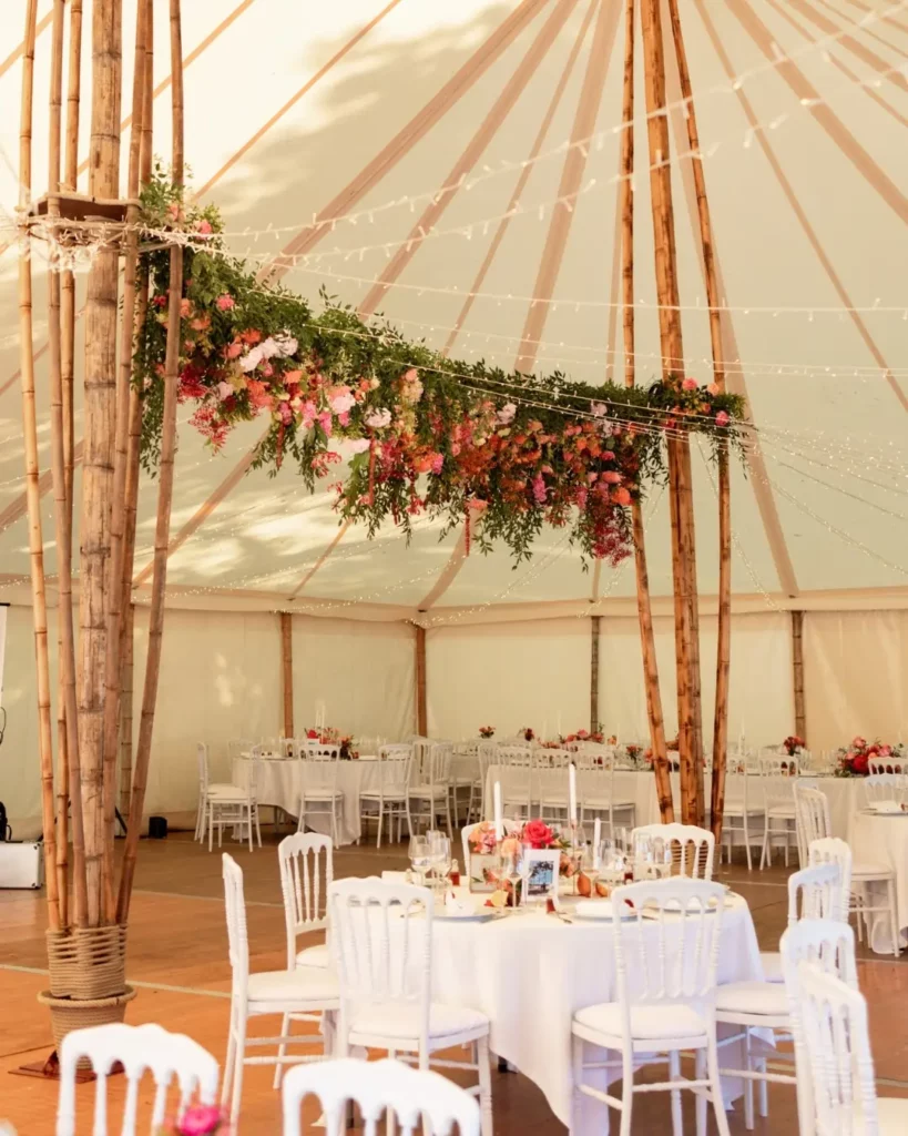 Décoration de mariage sous tente avec structure en bambou, guirlandes lumineuses et grand nuage de fleurs suspendu au-dessus des tables rondes.