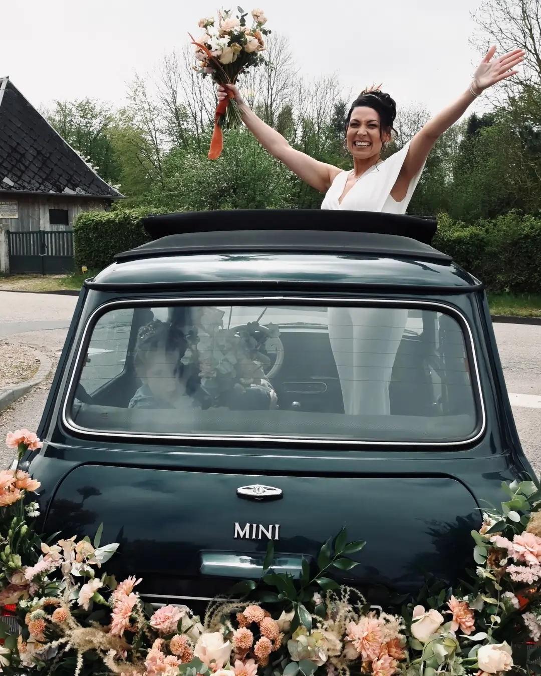 Mariée souriante saluant ses invités depuis une Mini décorée de fleurs pastel pour un accueil chaleureux.