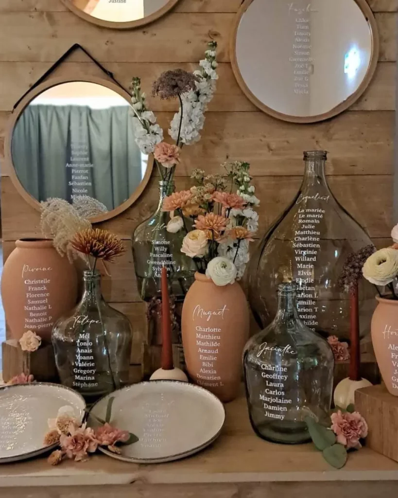 Plan de table en dame-jeanne et vases personnalisés, associé à une composition florale pour illustrer une scénographie de mariage créée avec de la location de décoration.