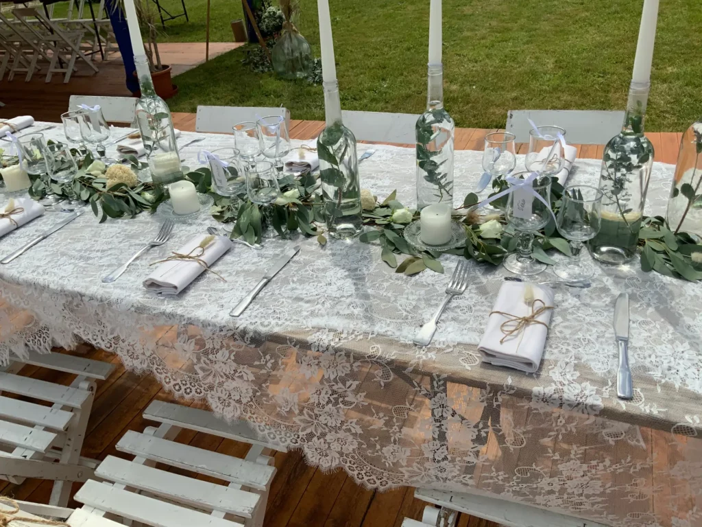 Table de mariage naturel chic avec nappe en dentelle, feuillage d’eucalyptus, bougies blanches et bouteilles en centre de table dans un décor en plein air.