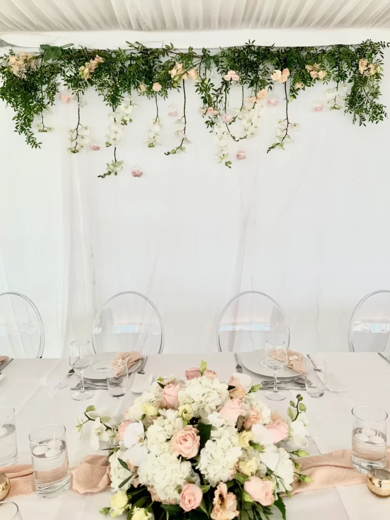 Chemin de table nude et composition florale blanche et rosée sur une table de mariage élégante, sous une cascade de fleurs suspendues.