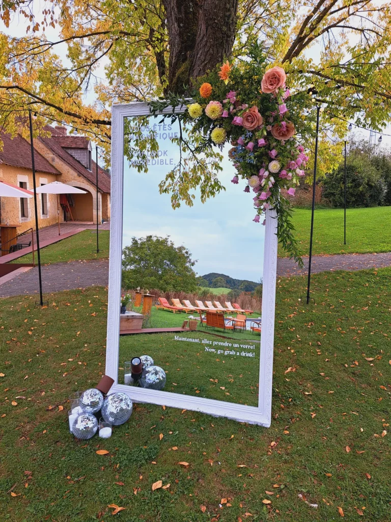 Grand miroir décoré de fleurs pour l’accueil des invités d’un mariage, installé en extérieur sous un arbre avec boules disco au sol.