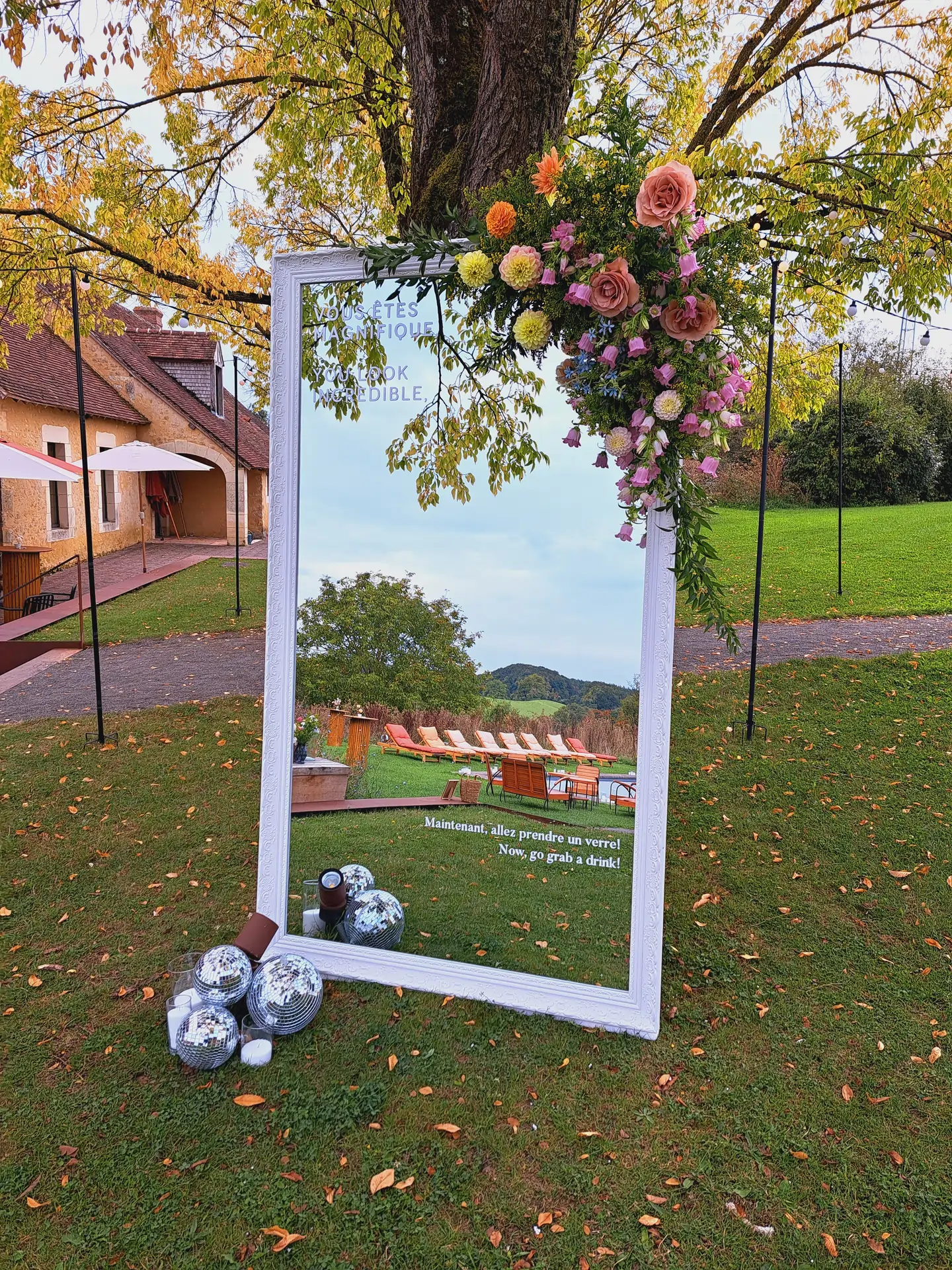 Location de miroirs pour mariage en Normandie. Grand miroir décoré de fleurs pour l’accueil des invités d’un mariage, installé en extérieur sous un arbre avec boules disco au sol.