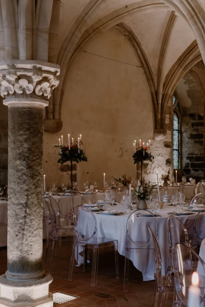 Salle de réception avec tables rondes et chaises transparentes, illustrant un choix de mobilier de mariage moderne dans un lieu en pierre.
