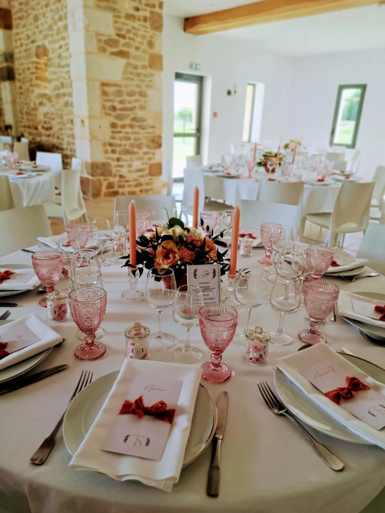 Table de mariage de printemps dressée dans une salle en pierre, avec verres roses, bougies pêche et centre de table fleuri aux tons plus vifs.