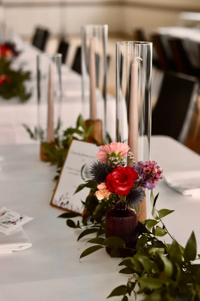 Centre de table simple et élégant pour un petit budget déco mariage, composé de feuillages, d’un mini vase coloré avec fleurs fraîches et de chandeliers en verre.