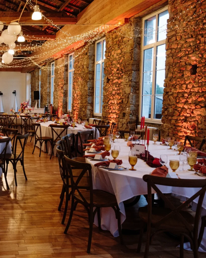 Décoration de réception au domaine de la Guérie en Normandie, avec tables dressées, guirlandes lumineuses au plafond, éclairages chauds sur les murs en pierre et art de la table raffiné. Décor floral par Maison Magnolia, photo d’Audrey Guyon.