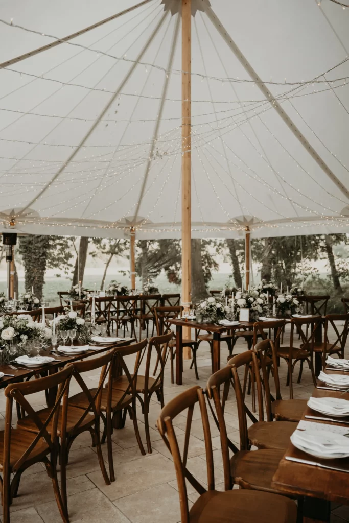 Décoration de mariage sous tente à La Maison Villeneuve, avec tables en bois, compositions florales blanches et guirlandes lumineuses suspendues, illustrant une mise en scène cohérente à l’opposé des erreurs fréquentes en décoration de mariage.