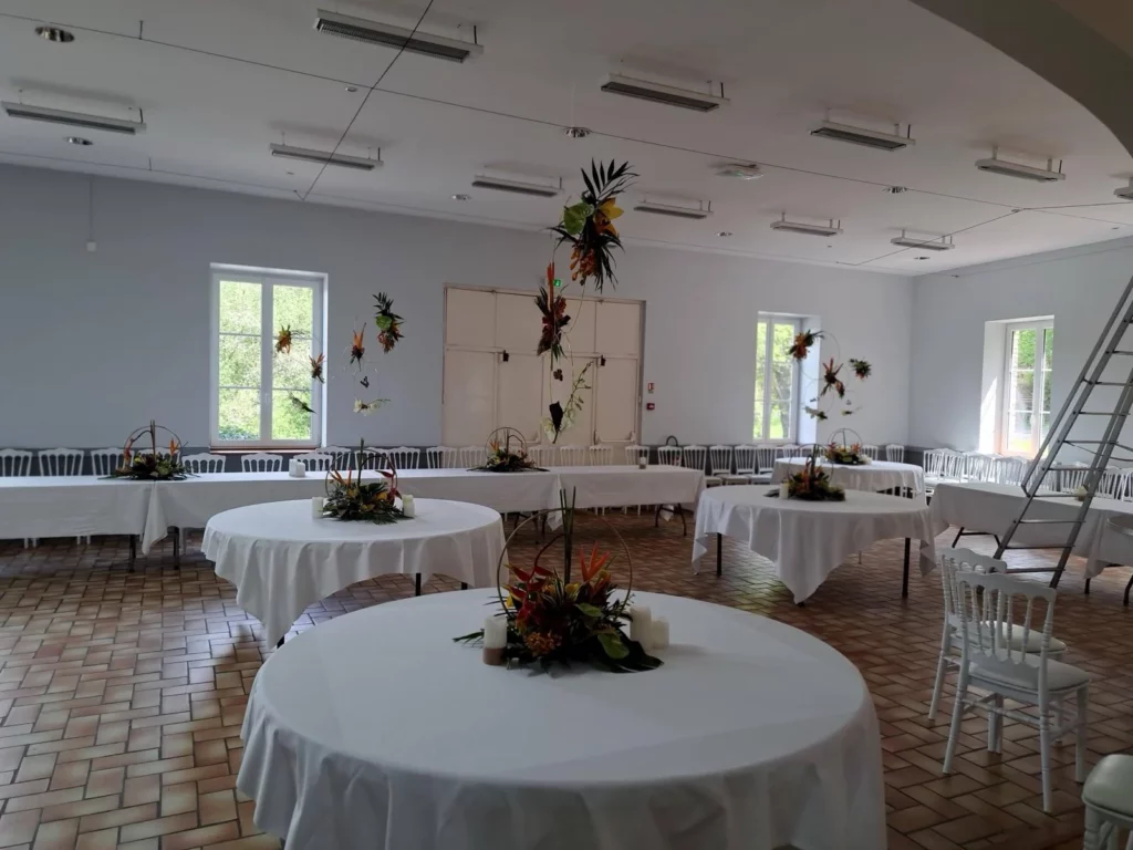 Salle de réception en cours d'installation, tables rondes nappées et cerceaux fleuris suspendus sous des néons La même idée transposée dans une salle réelle : néons au plafond, carrelage au sol, escabeau encore sorti.